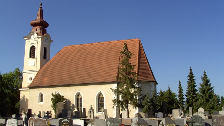 Außenansicht Kirche, © Johann Merl / Pfarrkirche Ebreichsdorf Außenansicht einer Kirche mit Friedhof im Vordergrund und blauem Himmel.