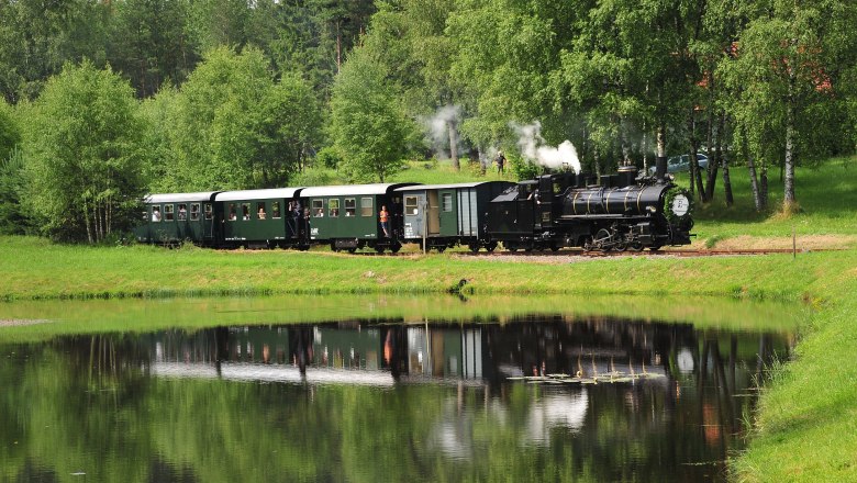 Waldviertelbahn, © knipserl.at Eine historische Dampflokomotive zieht grüne Waggons durch eine grüne Landschaft, die sich in einem ruhigen Teich spiegelt.