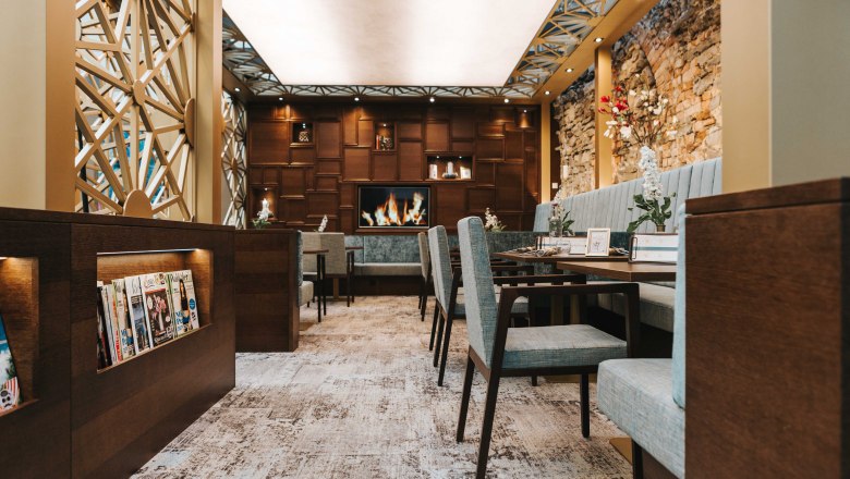 Café Wohnzimmer, © FMS Steinertor Gastronomie BetriebsgmbH Innenansicht eines stilvollen Cafés mit gemütlichen Sitzgelegenheiten, Kamin und Zeitschriftenregal.