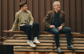 Weingut Fritz, © Weingut Fritz Zwei Männer sitzen entspannt auf gestapelten Holzbrettern vor einer dunklen Holzfassade und blicken zur Seite; ruhige, natürliche Atmosphäre in warmen Farbtönen.