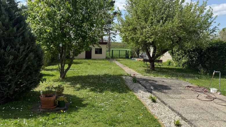 Garten, © Dana Perl Ein grüner Garten mit Bäumen, einem Weg und einem kleinen Gebäude im Hintergrund.