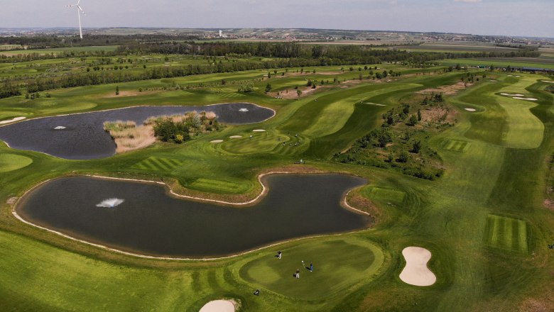 GolfRange in Bockfließ, © GEPA Pictures Luftaufnahme eines Golfplatzes mit Teichen und Windrad im Hintergrund.