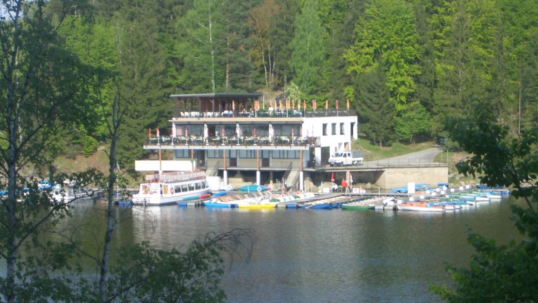 Bootsbetrieb und Seerestaurant, © MG Rfd Ein Seerestaurant mit Terrasse und Bootsanleger vor einem Wald.