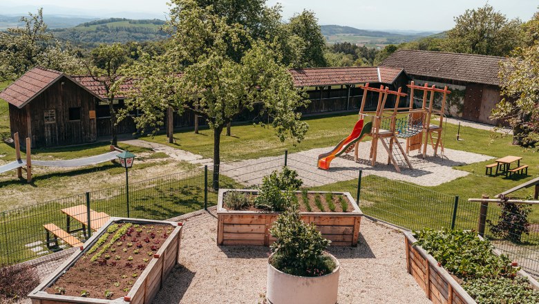 Wirtshaus mit Gast- und Nutzgarten, © Niederösterreich Werbung/Daniela Führer Ein Spielplatz mit Rutsche und Klettergerüst neben Hochbeeten in einem Garten mit Holzhütten im Hintergrund.