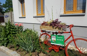 Weinbauernhof Wiesinger, © Weinbau Wiesinger Ein rotes Fahrrad mit Blumenkasten steht vor einem Gebäude mit Garten.