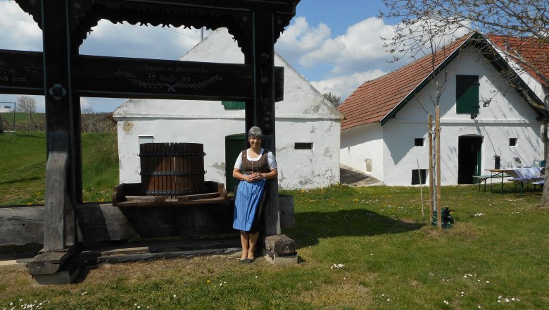 Kellergasse Sierndorf/March, © Franz Rauscher Eine Frau in traditioneller Kleidung steht neben einer alten Weinpresse vor einem historischen Gebäude mit weißen Wänden und roten Dächern.