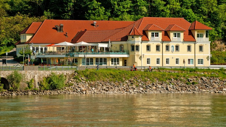 Residenz Wachau, © m+m webeagentur Ein gelbes Gebäude mit roten Dächern am Flussufer, umgeben von Bäumen.