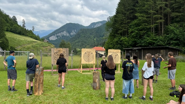 Wildnis Camps Wildniss Zentrum Nasswald, © Georg Bergthaler Gruppe von Menschen beim Axtwerfen im Freien vor Zielscheiben in einer bergigen Landschaft.