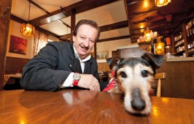 Landgasthaus Riefenthaler, © Franz Riefenthaler Ein Mann in einem Gasthaus lächelt, während ein Hund mit rotem Halstuch auf dem Tisch liegt.