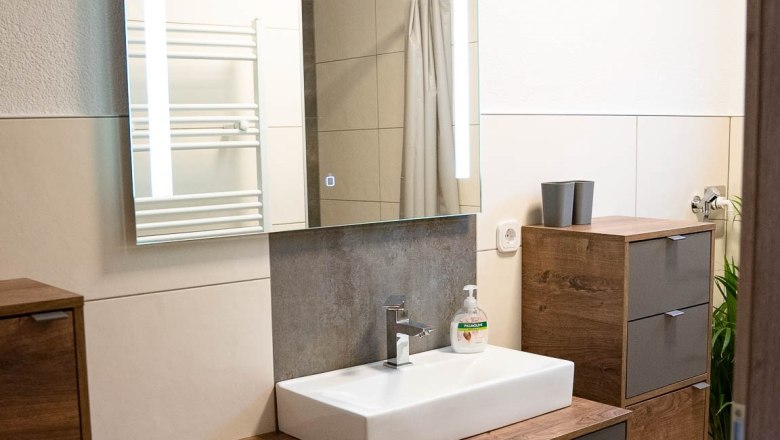 Badezimmer mit Dusche, © Manuel Hahn Modernes Badezimmer mit Holzschränken, Waschbecken und Spiegel.