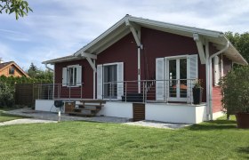 Haus gartenseitig, © Familie Mokesch Ein rotes Holzhaus mit weißem Dach und Fensterläden, umgeben von einem gepflegten Garten.