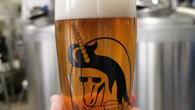 TDR-Bierglas design, © Drunken Unicorn Beer Factory Ein Bierglas mit Einhorn-Logo und der Aufschrift 'Tradition und Vielfalt' vor Brauereiausrüstung.