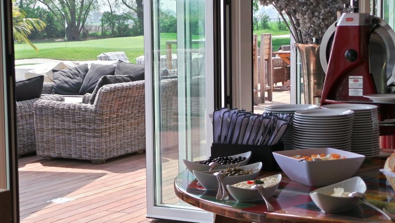 Boathouse, © DCC Ein gedeckter Tisch mit Geschirr und Snacks, daneben eine Terrasse mit Rattanmöbeln und Blick auf einen grünen Rasen.