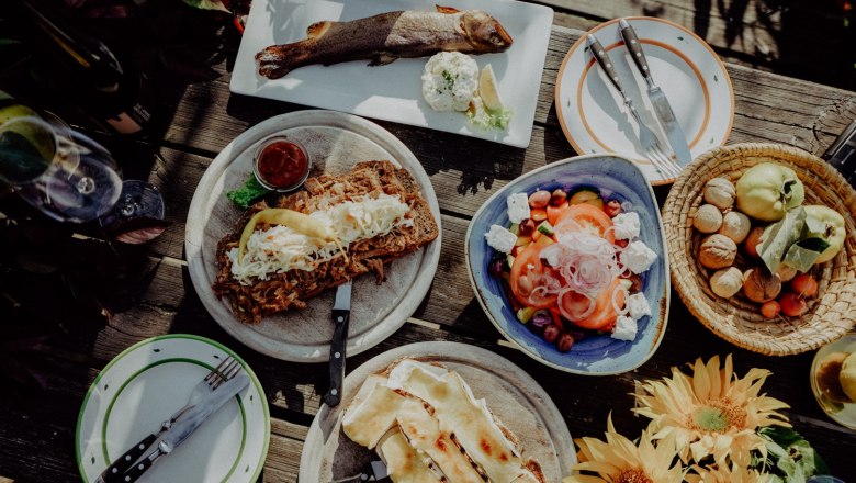 Auszug aus der Speisekarte, © Alter Klosterkeller Verschiedene Gerichte auf einem Holztisch, darunter ein ganzer Fisch, Salat, belegtes Brot und Sonnenblumen.