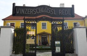 Winzerschlössl, © Weingut Redl Eingangstor zum Winzerschlössl Redl mit gelbem Gebäude im Hintergrund.