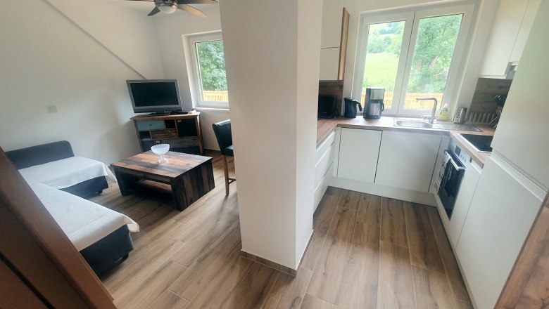 Wohnküche, © Tobias Micke Moderne Wohnküche mit Holzfußboden, Sofa, Fernseher, Couchtisch und weißer Einbauküche mit Fenster.