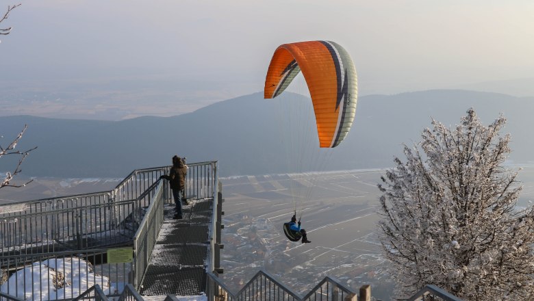 Naturpark Hohe Wand, © Naturpark Hohe Wand/Fraller Ein Paraglider fliegt über eine verschneite Landschaft im Naturpark Hohe Wand, während eine Person auf einer Aussichtsplattform steht.