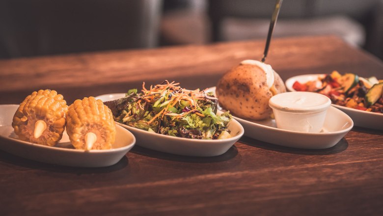 köstliche Beilagen, © Route 66 Fehr Verschiedene Beilagen auf einem Holztisch, darunter Maiskolben, Salat, Ofenkartoffel und Dip.