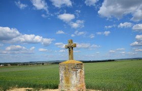 Marterl auf Oberer Leite, © Christian Häusler Ein steinernes Kreuz mit Christusfigur steht auf einem Feld unter blauem Himmel mit Wolken.