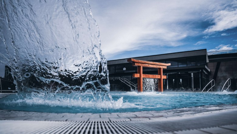 Linsberg Asia Therme, © Linbsberg Asia Ein Torii-Tor in einem Pool mit sprudelndem Wasser und einem modernen Gebäude im Hintergrund.