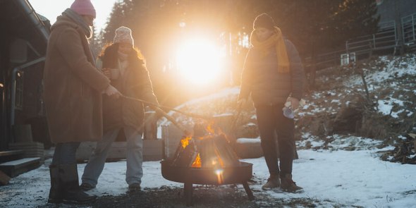 Wintermomente genießen, © Niederösterreich Werbung/friendship.is/Ian Ehm Drei Personen stehen rund um eine Feuerschale im Winter