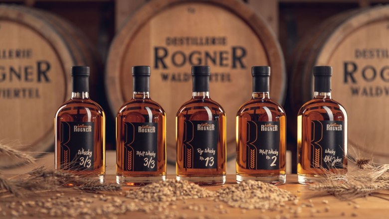 Sortiment Whisky, © Christoph Kerschbaum Fünf Whiskyflaschen der Destillerie Rogner vor Holzfässern.
