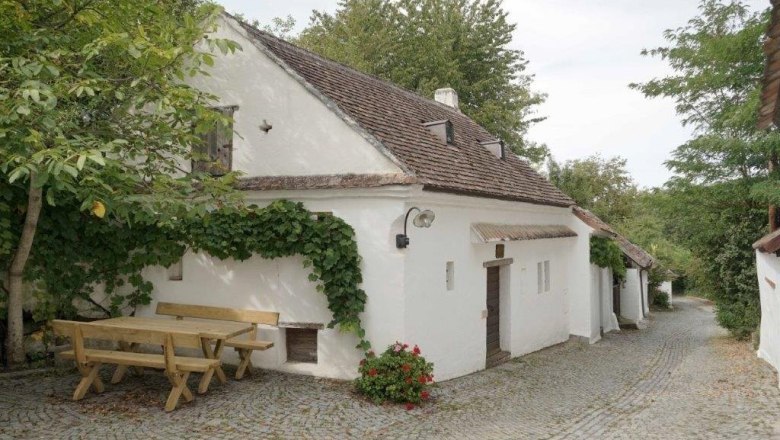 Weinviertler Kellergassenkulinarium, © D`Raschalaer Köllamauna Weißes Kellerstöckl mit Weinstöcken und Holztisch auf Kopfsteinpflasterstraße.