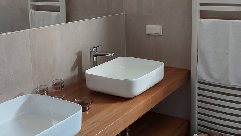 Badezimmer, © Bernadette Gierlinger Modernes Badezimmer mit zwei weißen Waschbecken auf Holzplatte, Handtuchhalter und Korb mit Handtüchern.