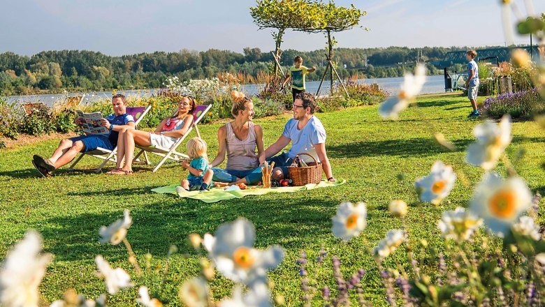 Gartenstadt Tulln, © Stadtgemeinde Tulln/Robert Herbst Familienpicknick im Grünen mit Blumen im Vordergrund und einem Fluss im Hintergrund.