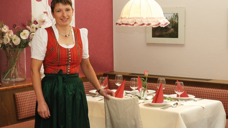 Gasthof Grossmann, © Gabriele Grossmann Eine Frau in traditioneller Kleidung steht neben einem gedeckten Tisch in einem Restaurant.