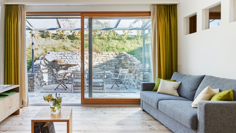 Weinviertler Kellerstöckl, © Andreas Hofer Innenansicht eines modernen Wohnzimmers mit grauem Sofa, Holzboden und großen Fenstern, die auf eine Terrasse mit Steinmauer und Gartenmöbeln führen.