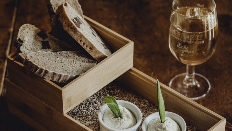 Selbstgebackenes Brot, © Niederösterreich Werbung/Sophie Menegaldo Ein Holztablett mit Brotscheiben, zwei Schälchen mit Aufstrich und ein Glas Weißwein auf einem Holztisch.