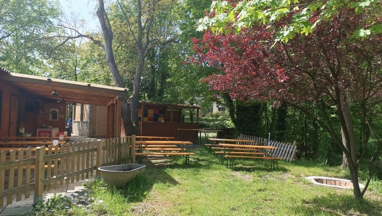 TDR-Zur MUCKE außen, © B.Grohotolszky Ein Biergarten mit Holzbänken und Tischen im Freien, umgeben von Bäumen und einem Holzzaun.