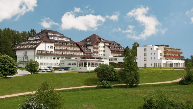 Hausansicht, © HKZGG, Robert Herbst Großes Gebäude mit mehreren Stockwerken und Balkonen, umgeben von grüner Landschaft und Bäumen, unter blauem Himmel.