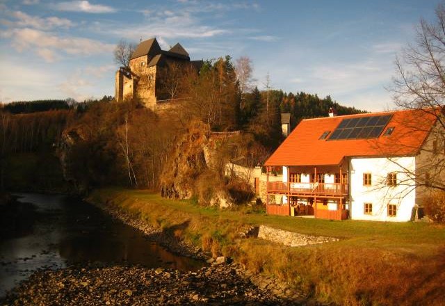Heurigen-Gasthof Prinz, © Dieter Prinz Ein Gasthof mit rotem Dach und Solarpanelen neben einem Fluss, im Hintergrund eine Burg auf einem Hügel.