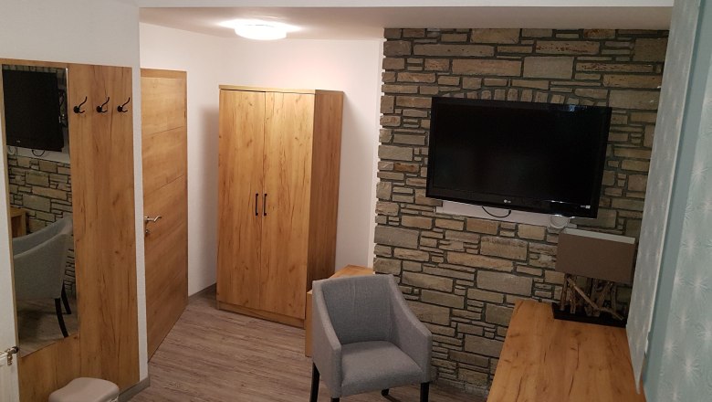 Pension Central, © Konrad Brandl Hotelzimmer mit Holzmöbeln, grauem Sessel und Flachbildfernseher an einer Steinwand.