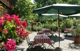 Gastgarten, © zVg Ein gemütlicher Gastgarten mit rot-weiß karierten Tischdecken, grünen Sonnenschirmen und blühenden Blumen im Vordergrund.