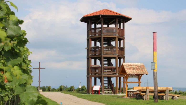 Aussichtsturm am Sandberg in Roseldorf, © Gemeinde Sitzendorf/ Schmida Ein hölzerner Aussichtsturm mit rotem Dach steht auf einem Hügel neben einem Kreuz und einer Sitzbank. Der Himmel ist bewölkt.