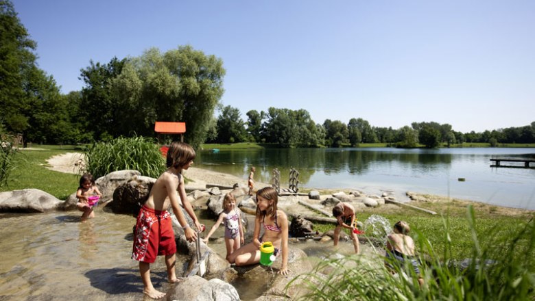 Aubad Tulln, © Stadtgemeinde Tulln, Alexander Haiden Kinder spielen im flachen Wasser eines Sees mit Steinen und Pflanzen im Hintergrund.