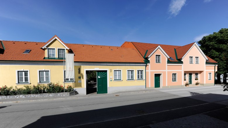 Haus, © Steve Haider Ein langes, einstöckiges Haus mit gelber und rosa Fassade, grünen Fensterläden und einem roten Ziegeldach unter blauem Himmel.