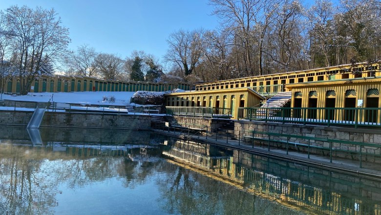 Nach der Sauna ins erfrischende Thermalwasser, © Wiener Alpen Außenansicht eines Thermalbads mit gelben Gebäuden und klarem Wasser, umgeben von schneebedeckten Bäumen.