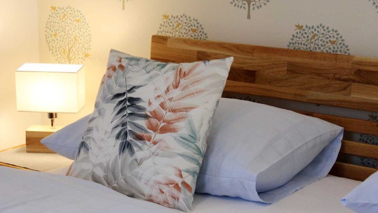 Gemütliche Zimmer, © Magdalena Donhauser Nahaufnahme eines Bettes mit Holzkopfteil, dekorativen Kissen und Nachttischlampe.