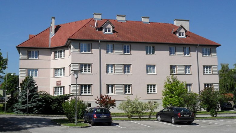 Schuberthof in Wimpassing, © frei Wohngebäude mit rotem Dach und mehreren Fenstern, umgeben von Bäumen und geparkten Autos.