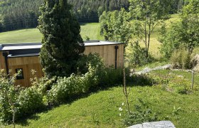 Tiny House - Hausansicht, © Stupbacher Ein modernes Holzhaus in einer grünen, hügeligen Landschaft mit Bäumen und Wiesen.