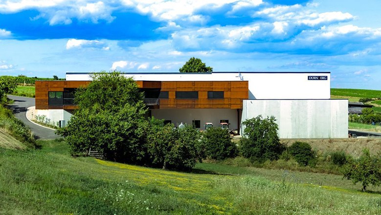 Weingut Dürnberg, © Weingut Dürnberg Moderne Weingut-Gebäude mit brauner und weißer Fassade, umgeben von grüner Landschaft und blauem Himmel.