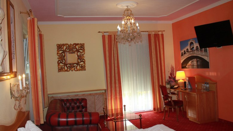 Suite, © Nimal Poudel Elegantes Hotelzimmer mit Kronleuchter, Ledersofa, Schreibtisch und großem Fenster mit Vorhängen.