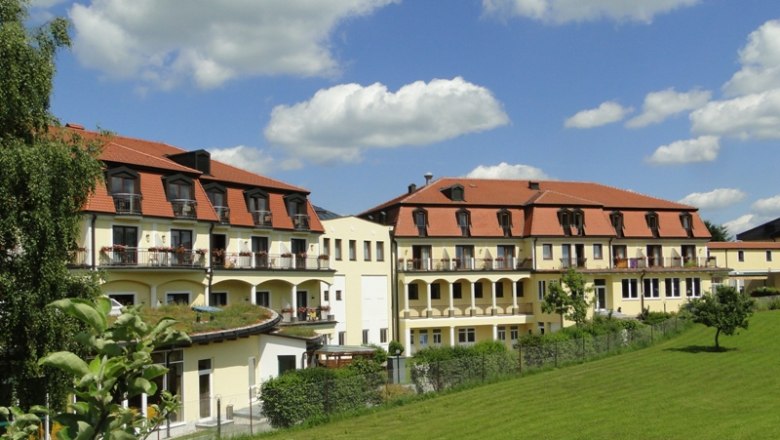 Kurhotel Moorbad Großpertholz, © Marktgemeinde Bad Großpertholz Kurhotel Moorbad Großpertholz mit rotem Dach und Balkonen, umgeben von grüner Wiese und Bäumen unter blauem Himmel.