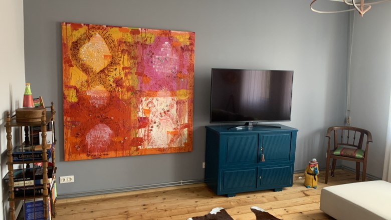 Stilvoll eingerichtet, © Fam. Wochesländer Wohnzimmer mit großem buntem Gemälde, Fernseher auf blauem Schrank, Holzboden und Kuhfell-Teppich.