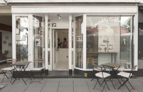 ²²Beans von außen, © Hönigmann/Roland Pohl Außenansicht eines Cafés mit Tischen und Stühlen auf dem Gehweg.