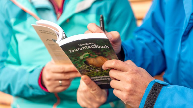 Lebensweg TourenTAGEbuch, © Lebensweg, Studio Kerschbaum Zwei Personen in blauen Jacken halten ein Buch mit dem Titel 'Lebensweg TourenTAGEbuch'.